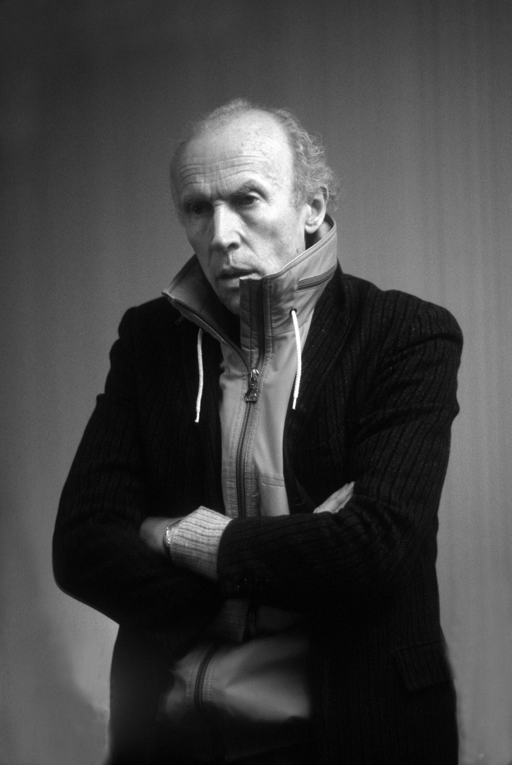 A figure stands with arms crossed, looking away from the camera with a pensive expression.