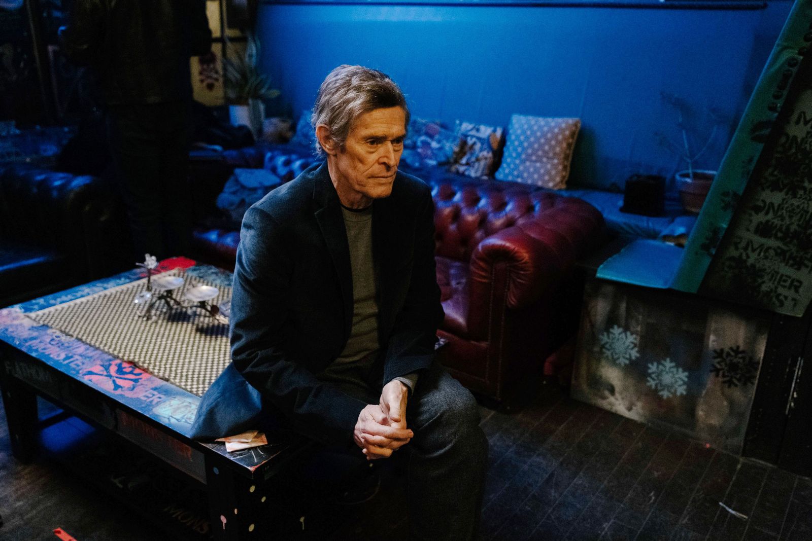 Willem Dafoe sitting on a table in blue room.